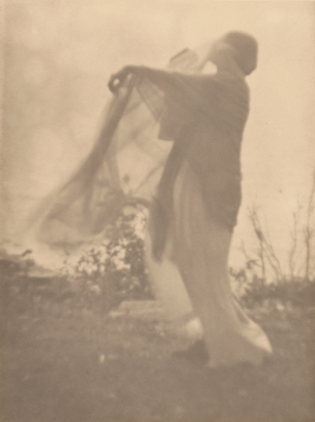 soft sepia photograph of a figure standing in a field, holding up their garments to the blowing wind