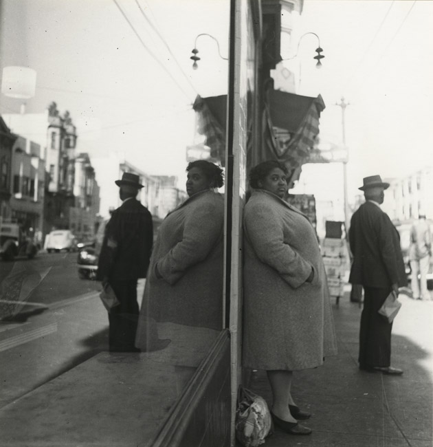 photograph of a woman standing next to a large shop window on the street, with her reflection doubled behind her as she gazes toward the camera