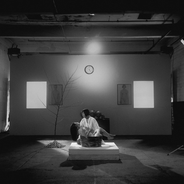 Photograph of a Japanese woman holding a girl in the center of a room on a floor cushion in the middle of the floor, between two windows and framed portraits, next to a small barren tree, 