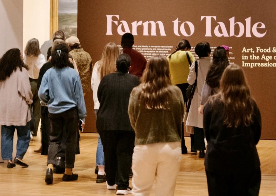 A group of people walk toward a brown wall display titled Farm to Table at an art exhibition, exploring how food-as-art is showcased. The floor is wooden, and the group appears to be viewing or entering the exhibit.